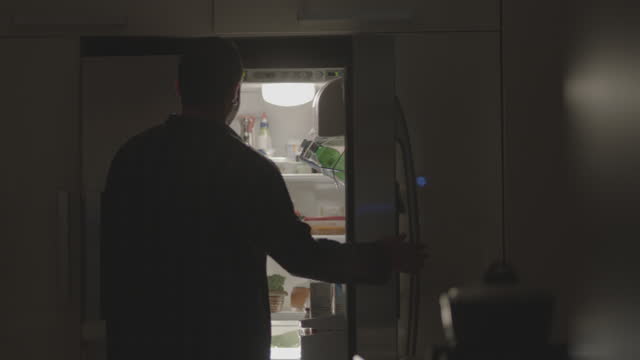 Young man opening the refrigerator at night, removing food, and closing it